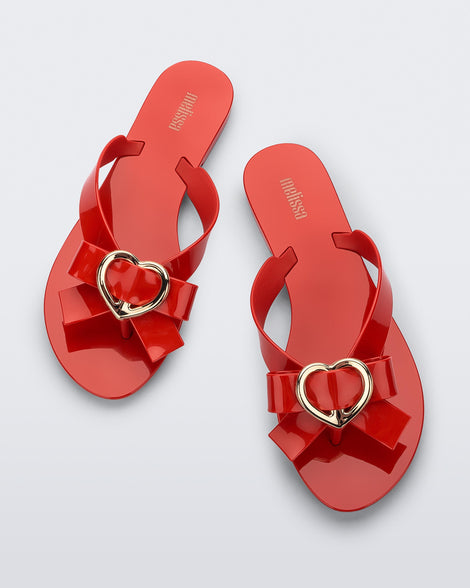Top view of a pair of red Melissa Harmonic Heart flip flops with a red bow and gold heart detail on the straps
