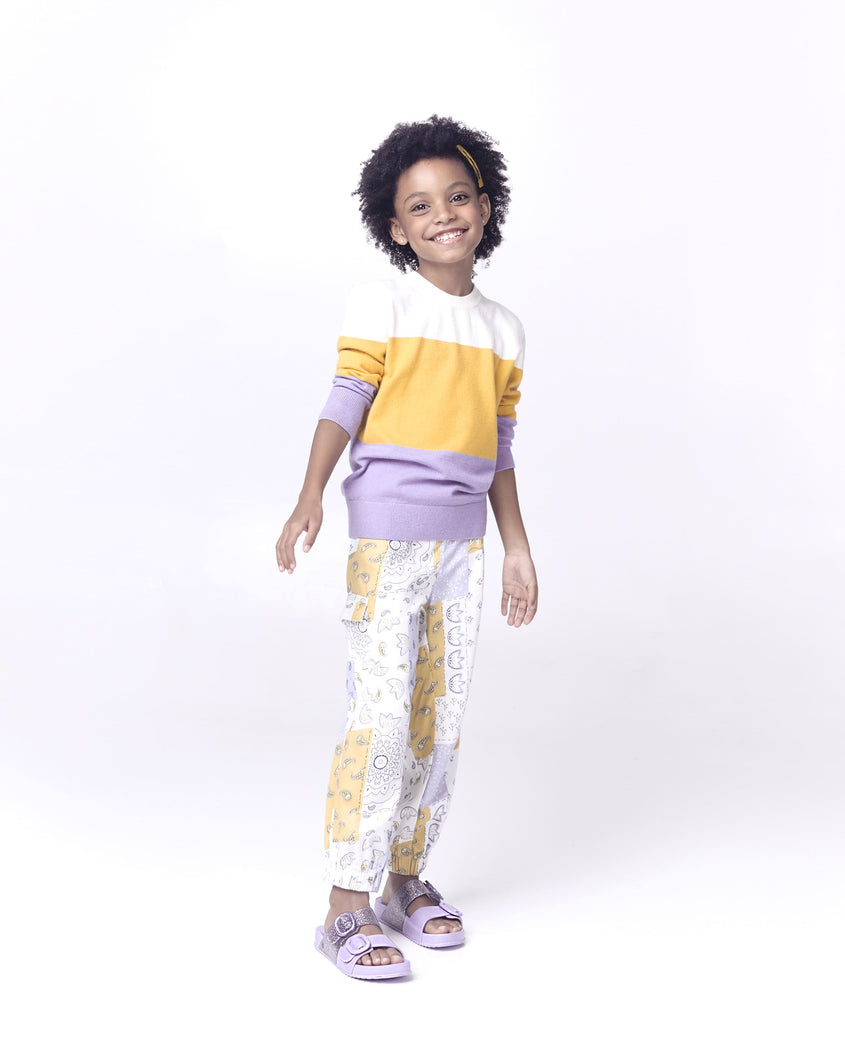 A kid model posing for a picture in a patterned top and bottom, wearing a pair of lilac Mini Melissa Cozy slides with two front straps with buckle details