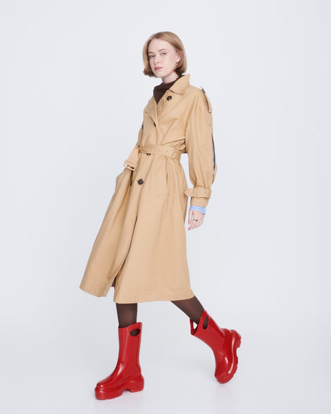 Model in a tan trench coat wearing a pair of red Ultra platform rain Boot with circle cutout on top