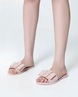 Model's legs  wearing a pair of light pink Babe slide sandals with large bow on top.