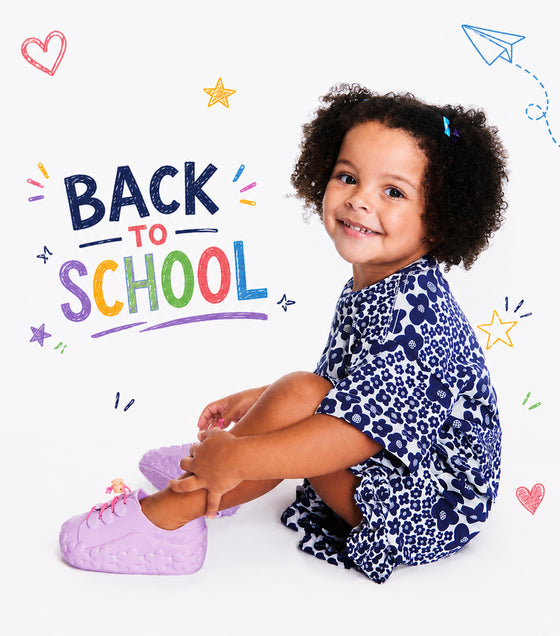 A young child with curly hair sits on a white background wearing a navy and white patterned dress and light purple sneakers. The image features a 'BACK TO SCHOOL' graphic with colorful doodles like stars, hearts, and paper airplanes.
