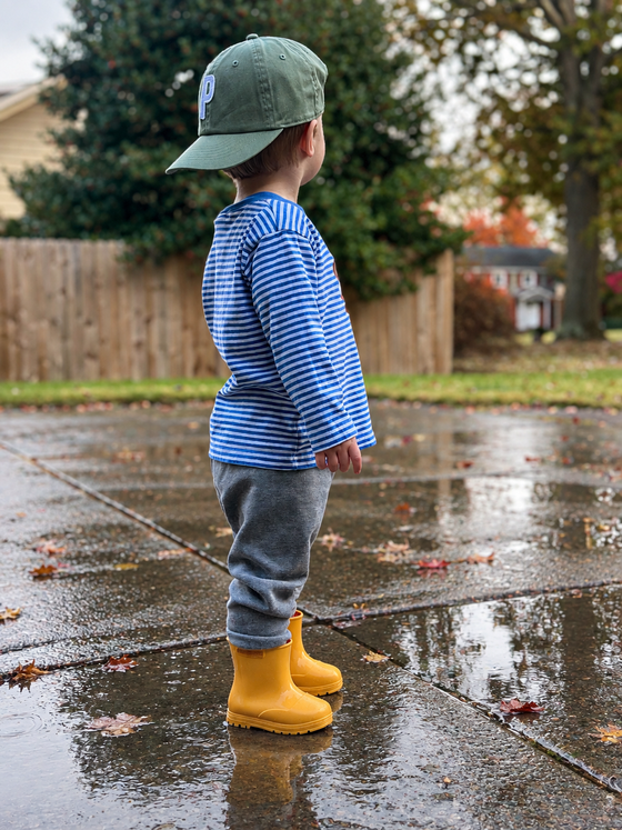 A toddler stands on a wet concrete driveway outdoors, wearing a blue and white striped long-sleeve shirt, gray pants, a green baseball cap, and bright yellow Welly rain boots.