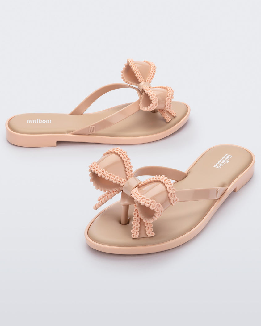 An angled front and side view of a pair of beige Melissa Slim flip flops with a lace like bow detail on the front strap.