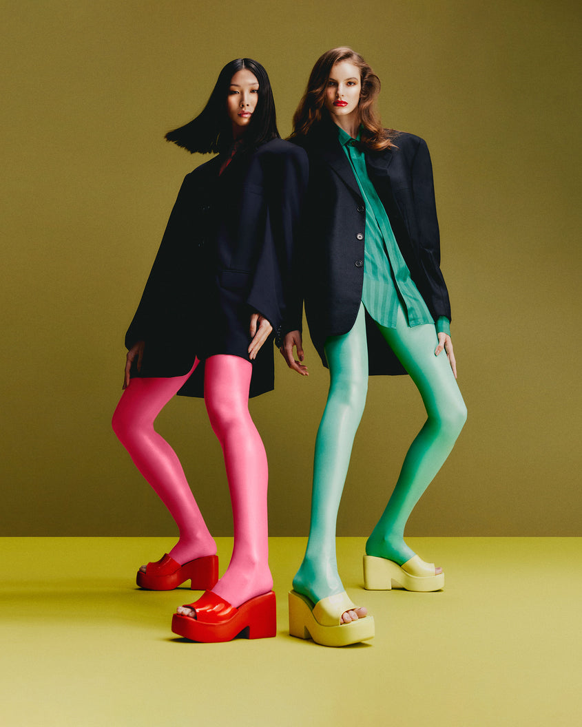 Two models in a standing pose each wearing a pair of Melissa Posh platform slides one pair is red and one pair is yellow
