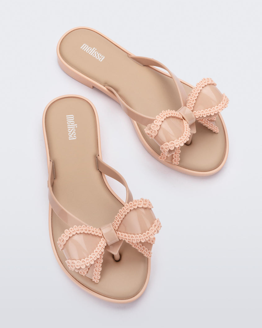 Top view of a pair of beige Melissa Slim flip flops with a lace like bow detail on the front strap.