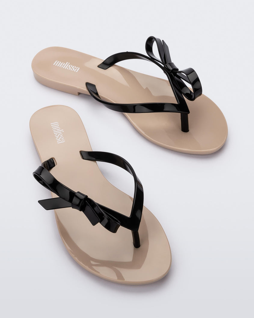 An front view of a pair of beige/black Melissa Harmonic Sweet flip flops with a beige sole and black straps with a bow detail on the front.