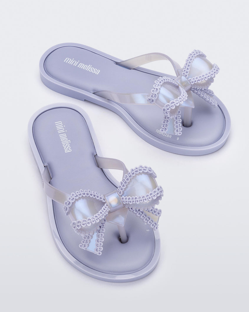 An angled front and top view of a pair of lilac Mini Melissa Slim flip flops with a lace like bow detail on the front strap.
