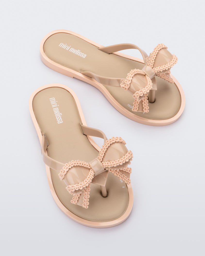 An angled top view of a pair of beige Mini Melissa Slim flip flops with a lace like bow detail.