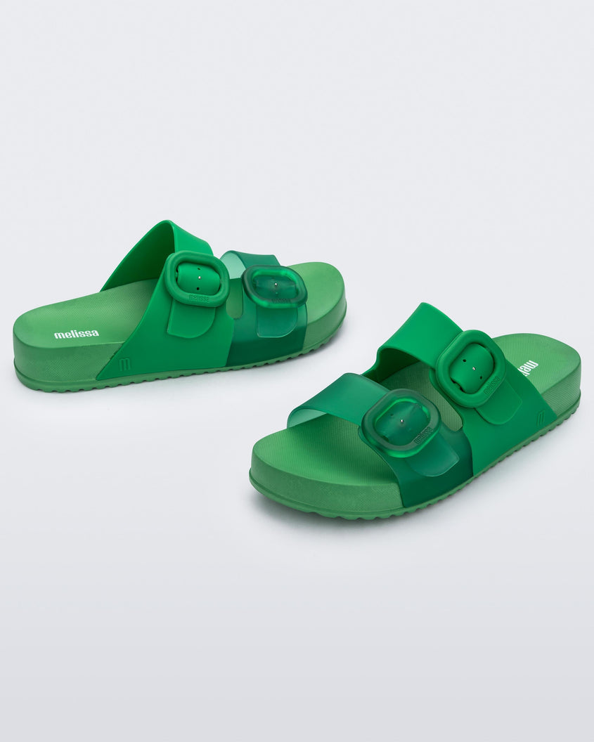 An angled front and side view of a pair of Green Melissa Cozy slides with two green and transparent green straps with a buckle detail.