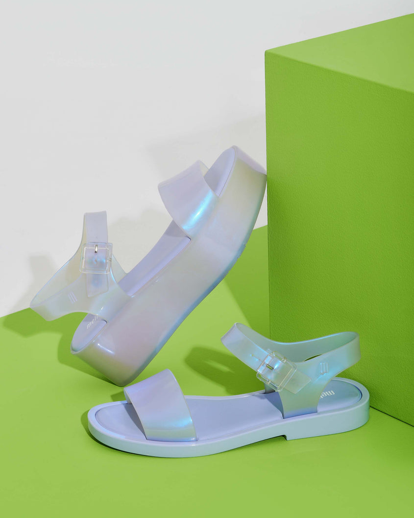 An angled view of a pearly blue Melissa Mar Platform sandal leaning on a green block next to a lilack/blue pearl Melissa Mar sandal sitting on a green surface.