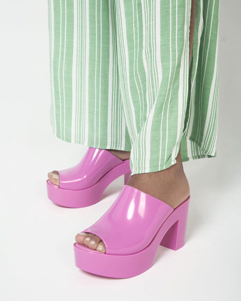 A model's legs wearing pink Melissa Mule platform heels and green pants.