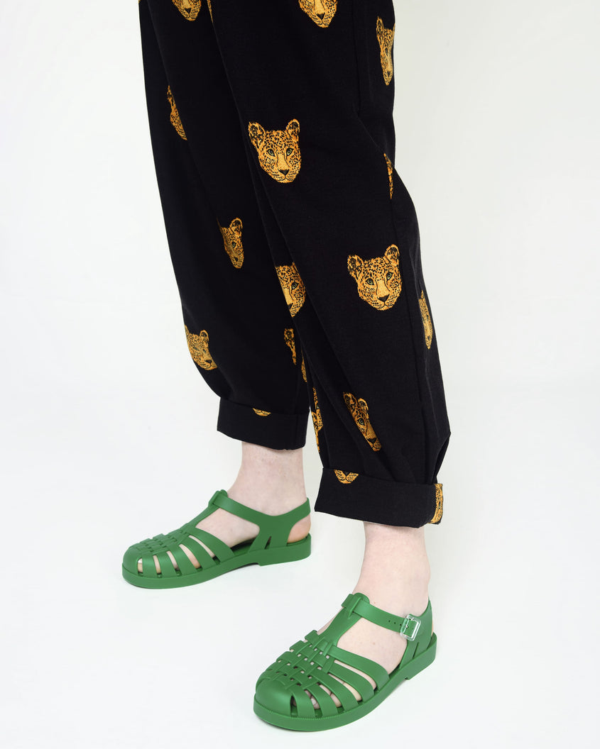 Model's legs in black/gold printed paints wearing a pair of green Melissa Possession sandals with a closed toe front weft design connected to a top strap with a buckle.