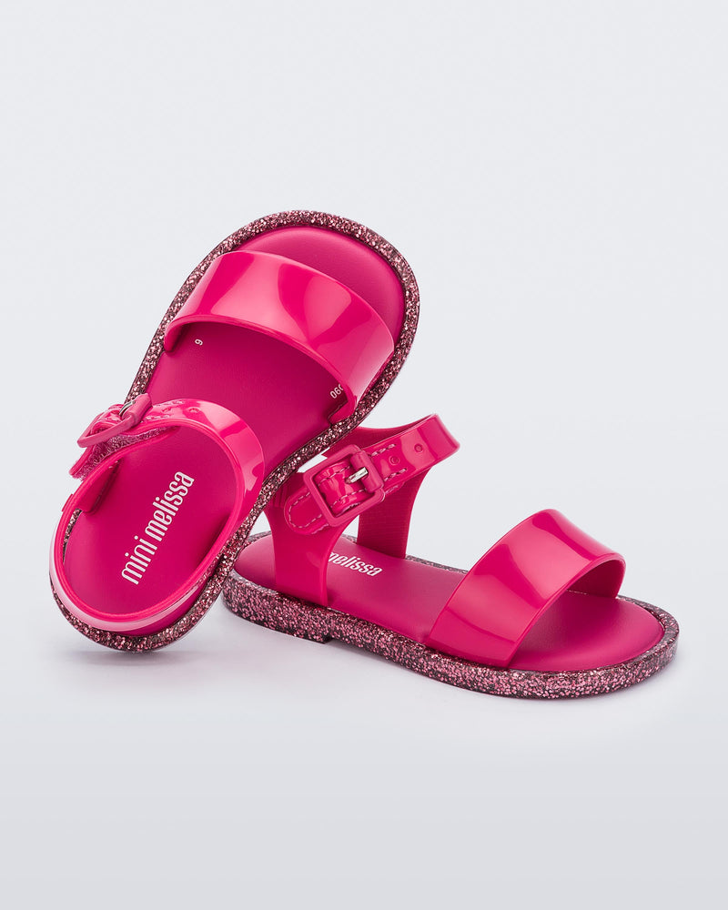 An angled top and side view of a pair of pink/pink glitter Mini Melissa sandal with two straps and a pink buckle, leaning on eachother.