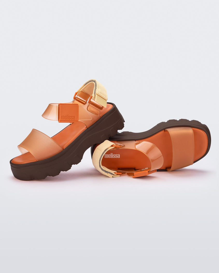 An angled top and side view of a pair of orange/clear orange Melissa platform Kick Off sandals with two straps and a brown sole.