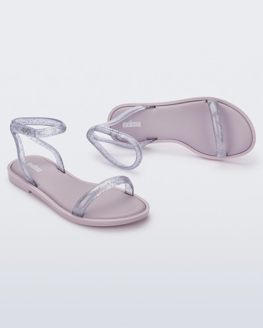 An angled front and side view of a pair of lilac/clear glitter Melissa Wave Sandals with a lilac base and a clear glitter front and ankle straps.
