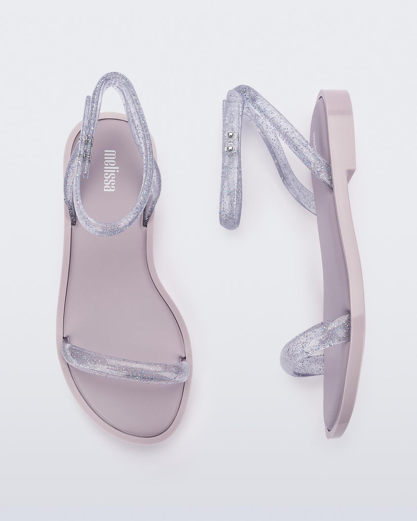 A top and side view of a pair of lilac/clear glitter Melissa Wave Sandals with a lilac base and a clear glitter front and ankle straps.