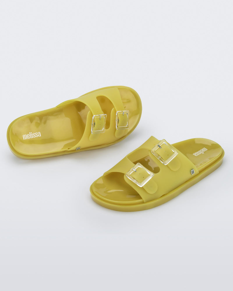 An angled front and top view of a pair of yellow Melissa Wide Slides with two straps fastened on top with two clear buckles and a "m" logo on the side.