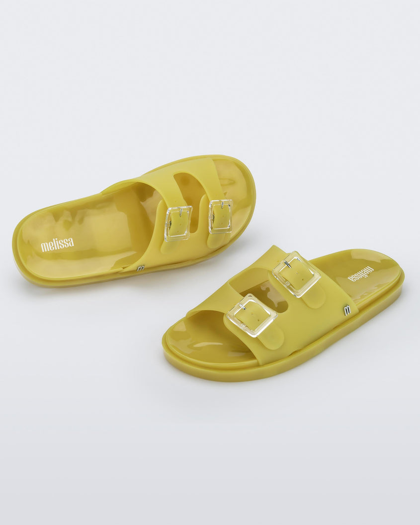 An angled front and top view of a pair of yellow Melissa Wide Slides with two straps fastened on top with two clear buckles and a 