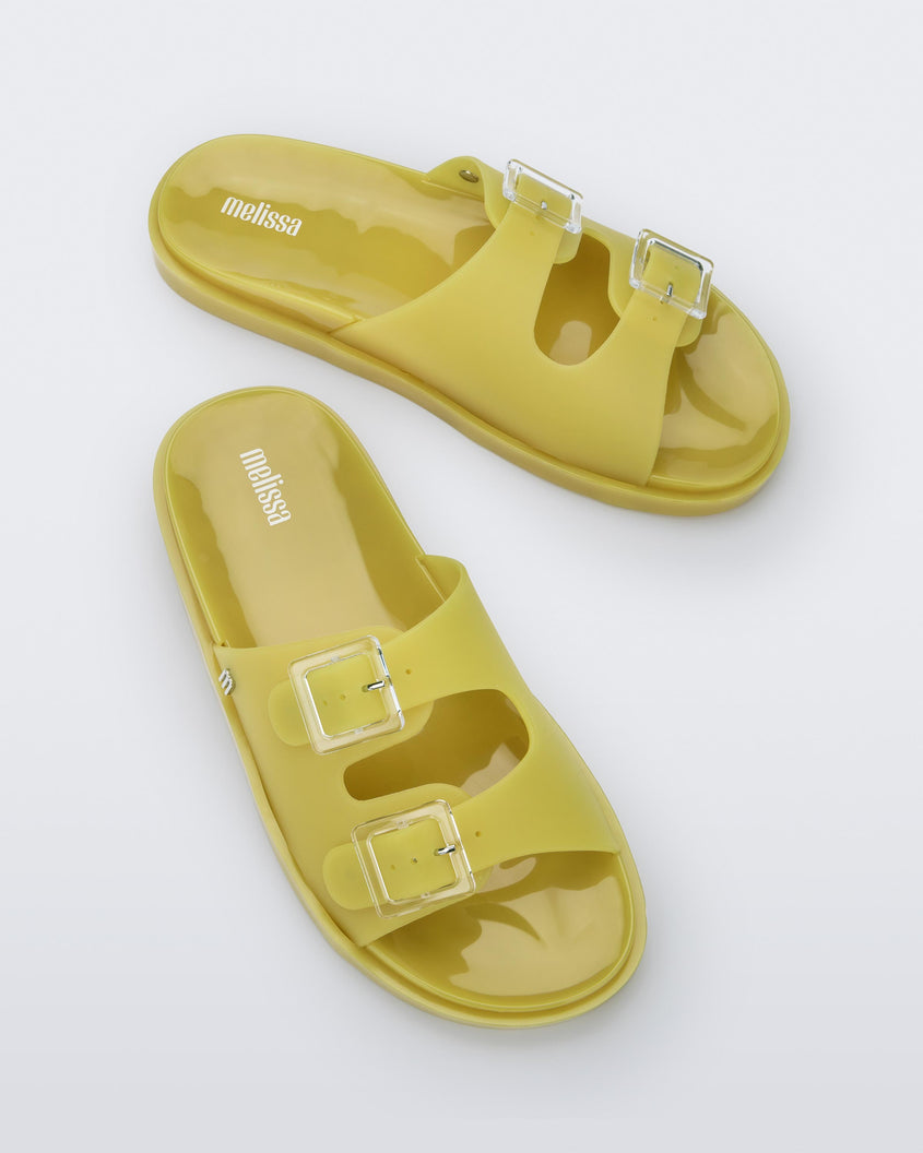 An angled top view of a pair of yellow Melissa Wide Slides with two straps fastened on top with two clear buckles and a 