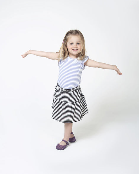 A child model posing in a striped skirt and tshirt wearing a pair of Mini Melissa Campana flats in purple glitter with an open woven texture.