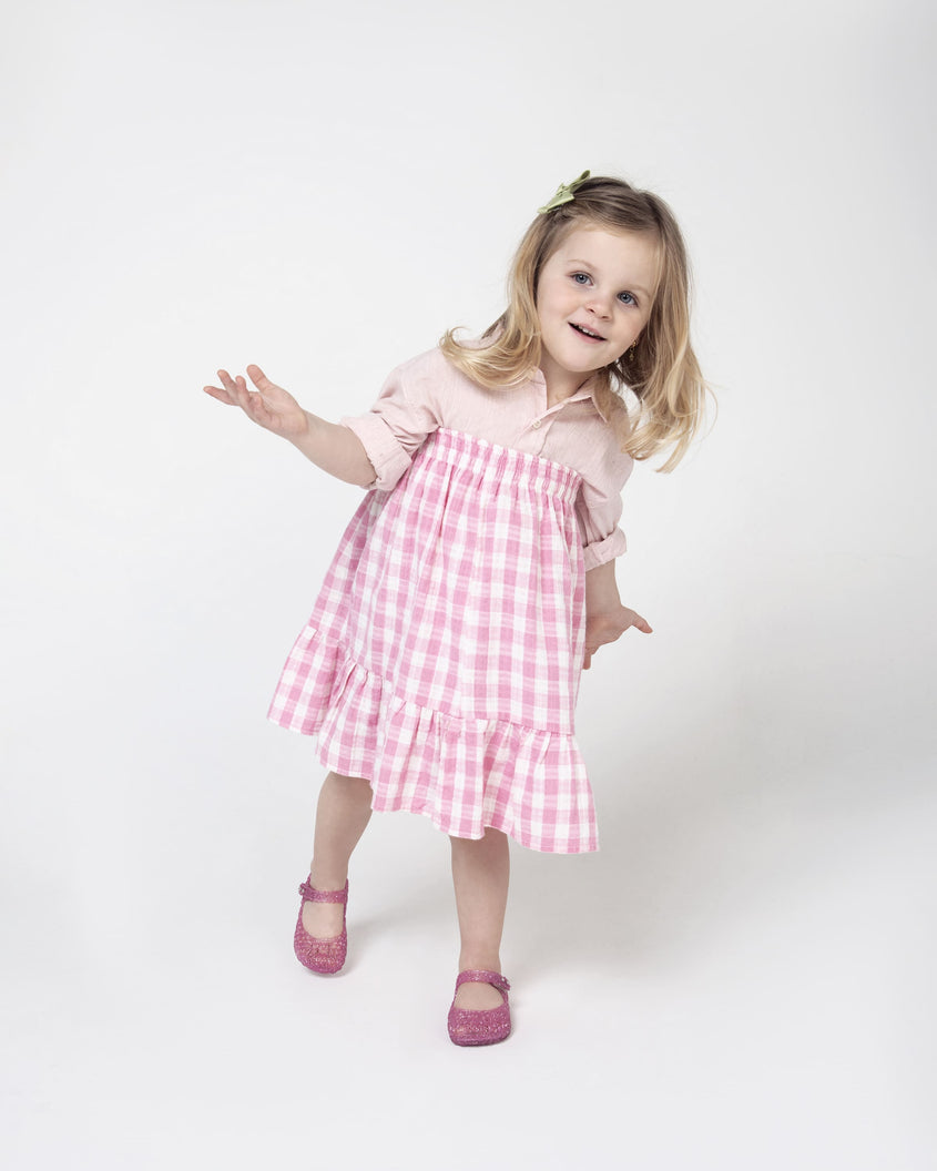 A child model in a beige top and a pink gingham skirt, wearing a pair of pink glitter Mini Melissa Campana flats with a snap strap for baby and an open woven texture.