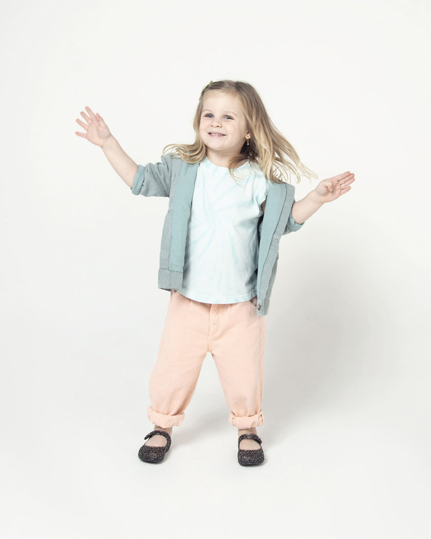 A child model posing in peach colored jeans with a white top wearing a pair of the Mini Melissa Campana flats for baby in black glitter with an open woven texture.
