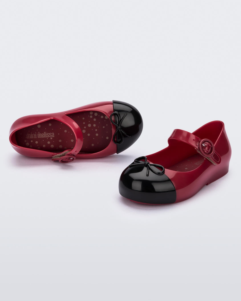An angled front and side view of a pair of Red/Black Mini Melissa Sweet Love Cap Toe flats with a red base, top strap and a black toe cap with a bow detail on top.