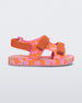 Side view of a orange/pink Mini Melissa Ping Pong sandal with a pink, orange and yellow floral patterned base and orange velcro straps.