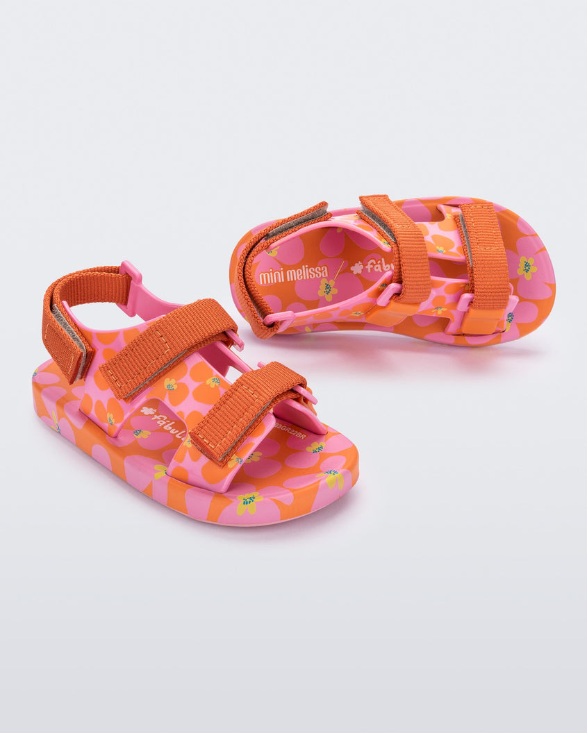 An angled side and top view of a pair of orange/pink Mini Melissa Ping Pong sandals with a pink, orange and yellow floral patterned base and orange velcro straps.