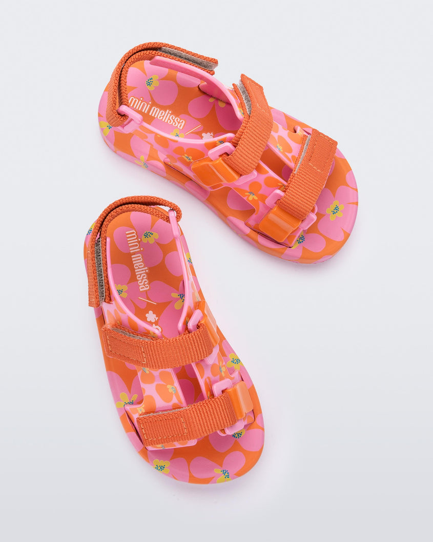 Top view of a pair of orange/pink Mini Melissa Ping Pong sandals with a pink, orange and yellow floral patterned base and orange velcro straps.