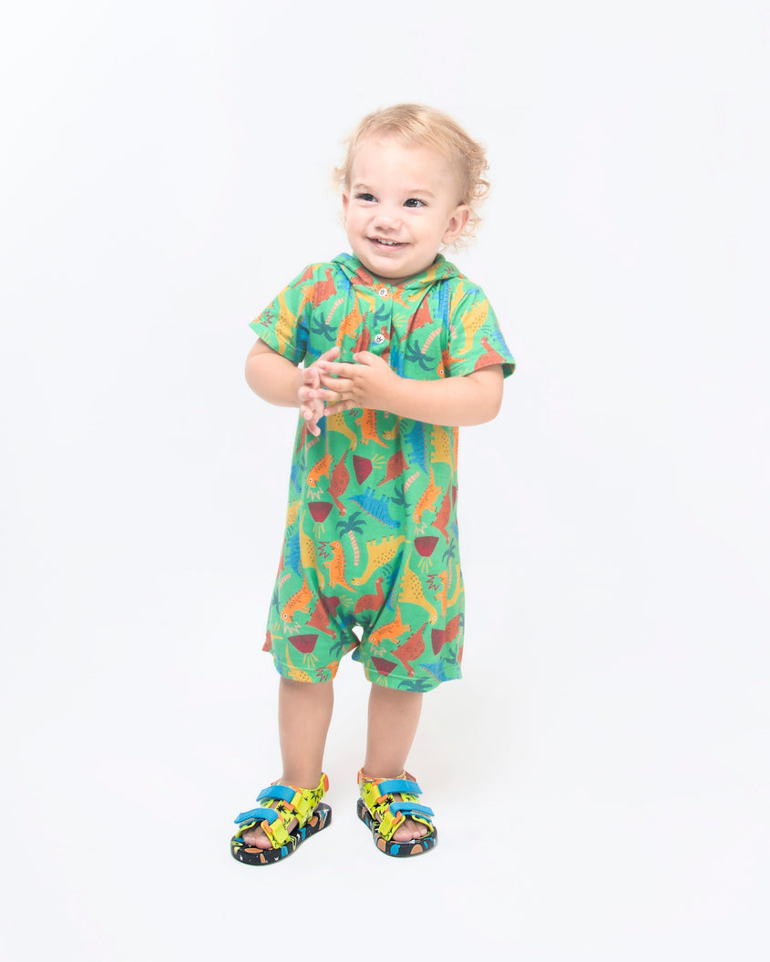 A young model in a green jumpsuit, wearing a pair of black/green Mini Melissa Ping Pong sandals with a green patterned base, black patterned sole, two blue front velcro straps and and an orange back velcro strap.