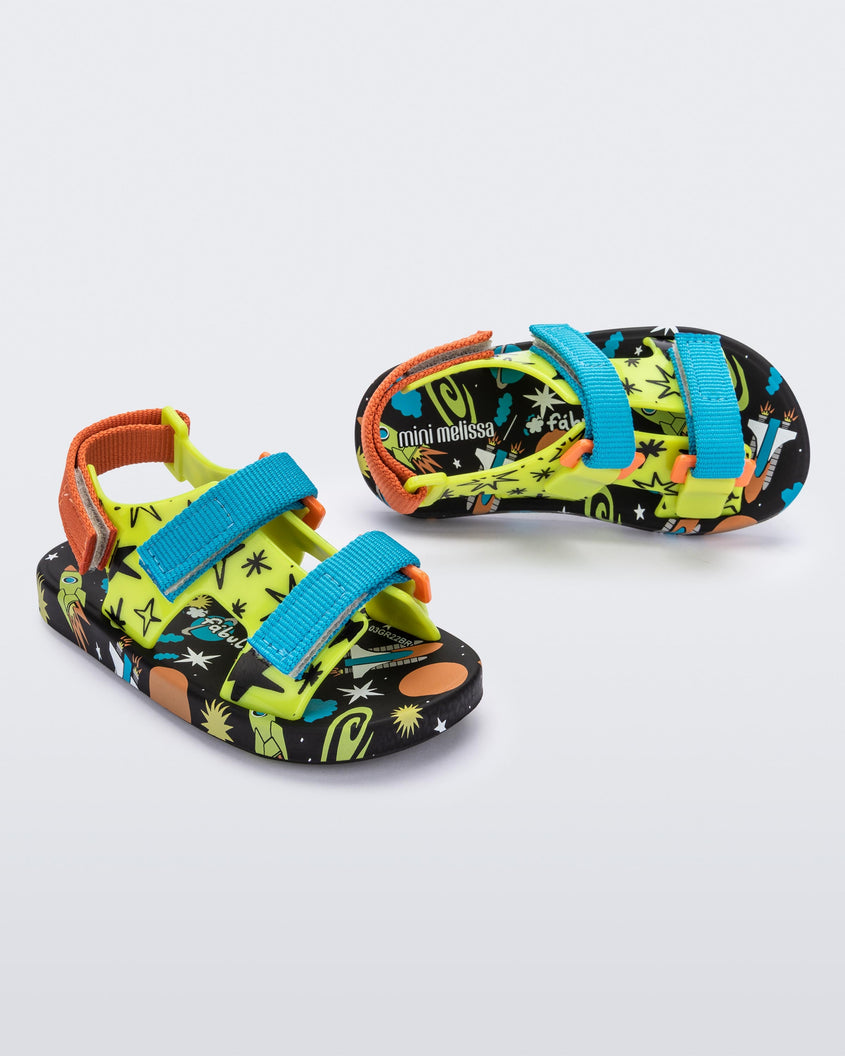 An angled side and top view of a pair of black/green Mini Melissa Ping Pong sandals with a green patterned base, black patterned sole, two blue front velcro straps and and an orange back velcro strap.