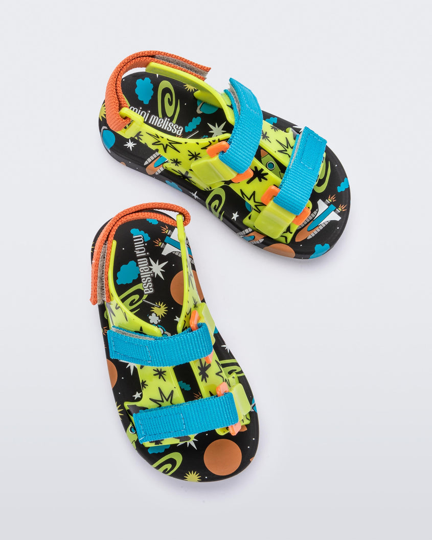 Top view of a pair of black/green Mini Melissa Ping Pong sandals with a green patterned base, black patterned sole, two blue front velcro straps and and an orange back velcro strap.