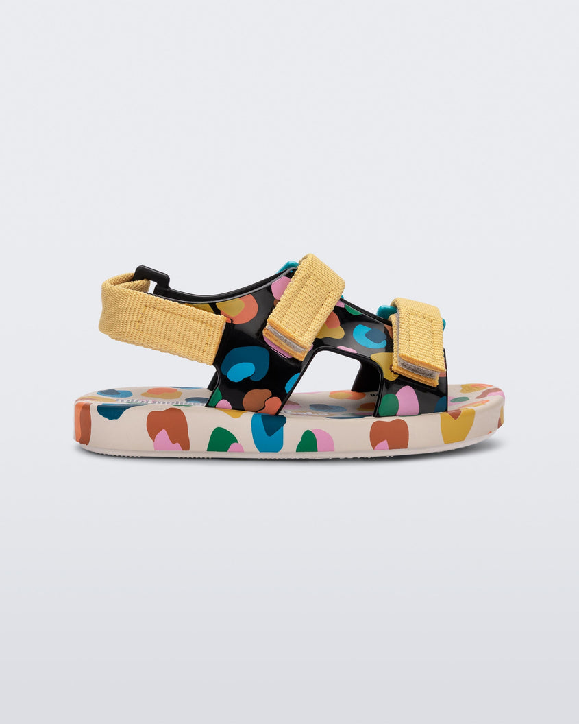 Side view of a beige/mustard Mini Melissa Ping Pong sandal with a black multicolored patterned base, white multicolored sole and yellow velcro straps.