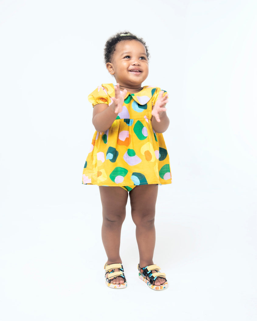 A baby model wearing a yellow patterned dress and a pair of beige/mustard Mini Melissa Ping Pong sandals with a black multicolored patterned base, white multicolored sole and yellow velcro straps.
