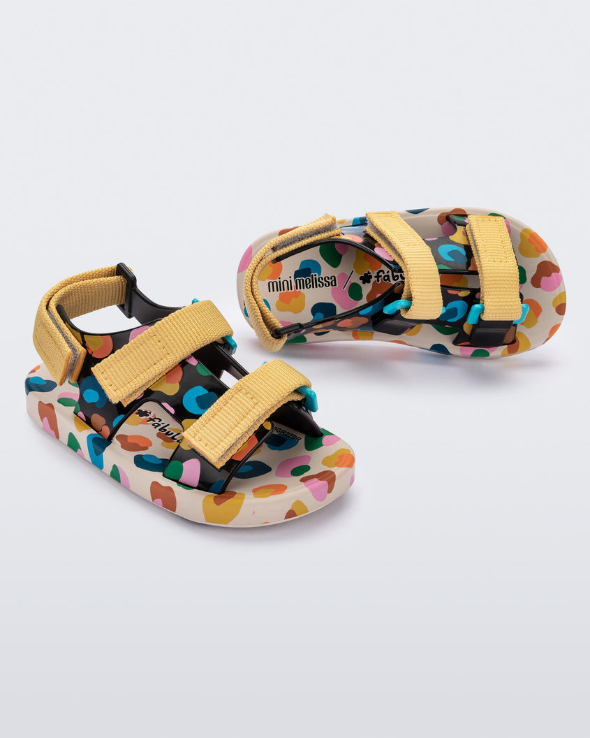 An angled side and top view of a pair of beige/mustard Mini Melissa Ping Pong sandals with a black multicolored patterned base, white multicolored sole and yellow velcro straps.