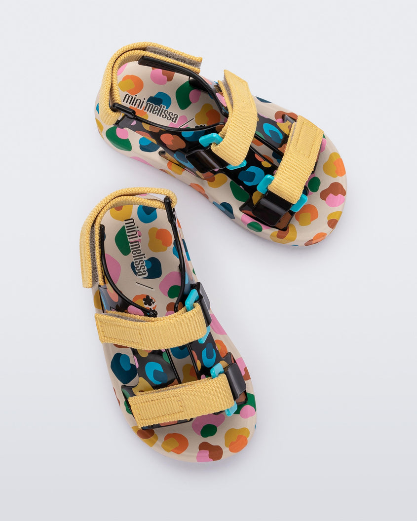 Top view of a pair of beige/mustard Mini Melissa Ping Pong sandals with a black multicolored patterned base, white multicolored sole and yellow velcro straps.