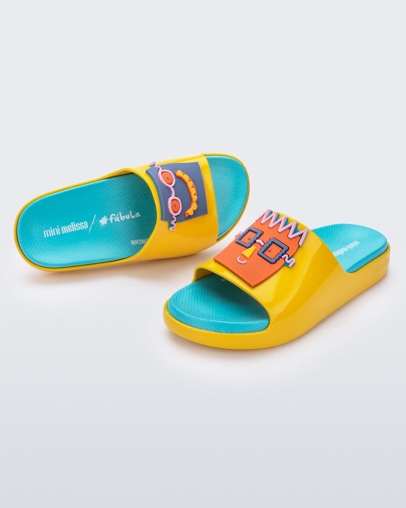 An angled top and side view of a pair of yellow Mini Melissa Cloud slides with a blue insole, yellow base and a cartoon drawn smiley face on the front.
