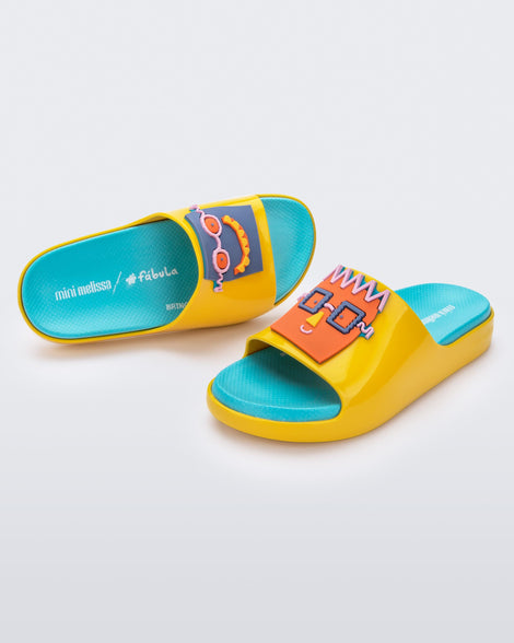 An angled top and side view of a pair of yellow Mini Melissa Cloud slides with a blue insole, yellow base and a cartoon drawn smiley face on the front.