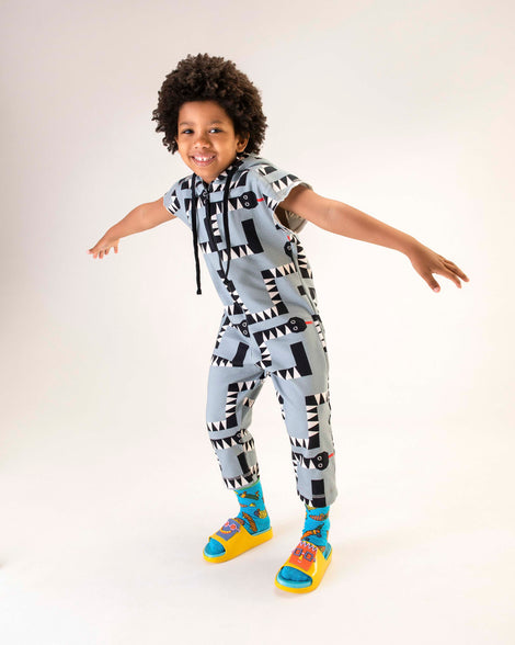 A model posing for a picture in a jumpsuit and a pair of yellow Mini Melissa Cloud slides with a blue insole, yellow straps and a cartoon drawn smiley face on the front strap.