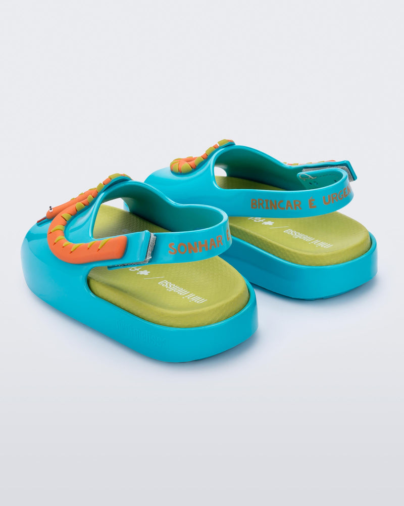 Angled view of the back of pair of blue/green Mini Melissa Cloud sandals with a velcro back strap, a green insole, blue base and orange snake detail on the front.