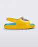 Side view of a pair of yellow Mini Melissa Cloud sandals with a velcro back strap, blue insole, yellow base and a cartoon drawn smiley face on the front.