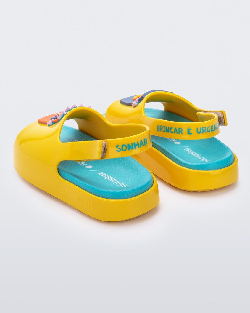 An angled back view of a pair of yellow Mini Melissa Cloud sandals with a velcro back strap, blue insole, yellow base and a cartoon drawn smiley face on the front.
