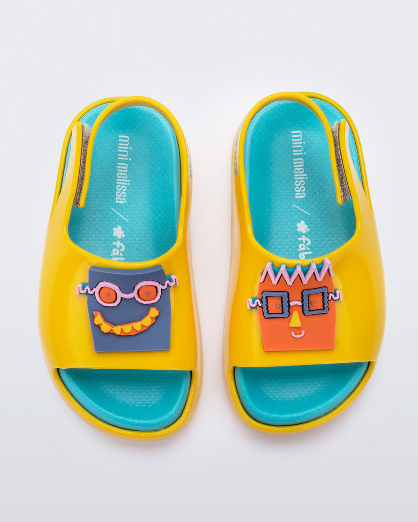 Top front view of a pair of yellow Mini Melissa Cloud sandals with a velcro back strap, blue insole, yellow base and a cartoon drawn smiley face on the front.
