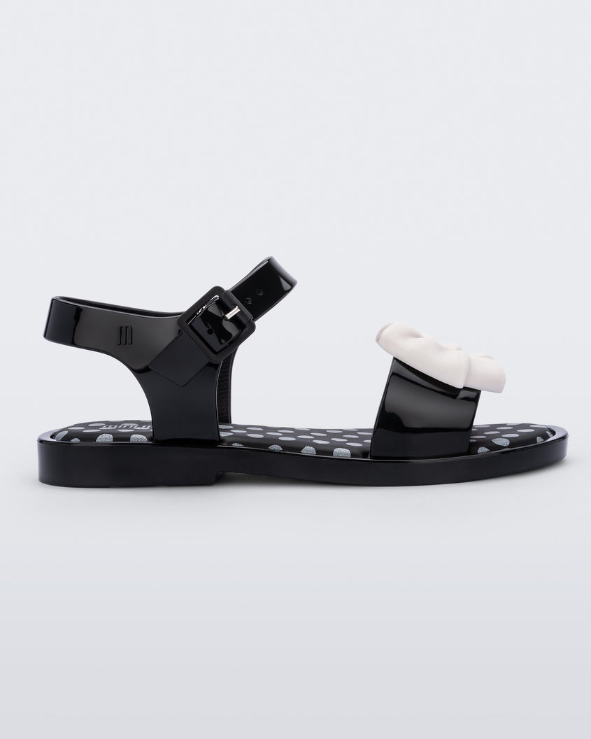 Side view of a black/beige Mini Melissa Mar Princess sandals with a silver polka dot design insole and a white glitter bow detail on the front strap.