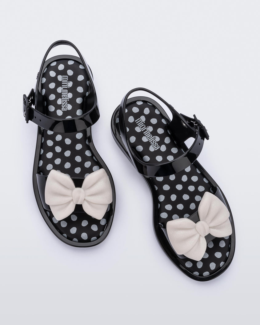 Top view of a pair of black/beige Mini Melissa Mar Princess sandals with a silver polka dot design insole and a white glitter bow detail on the front strap.