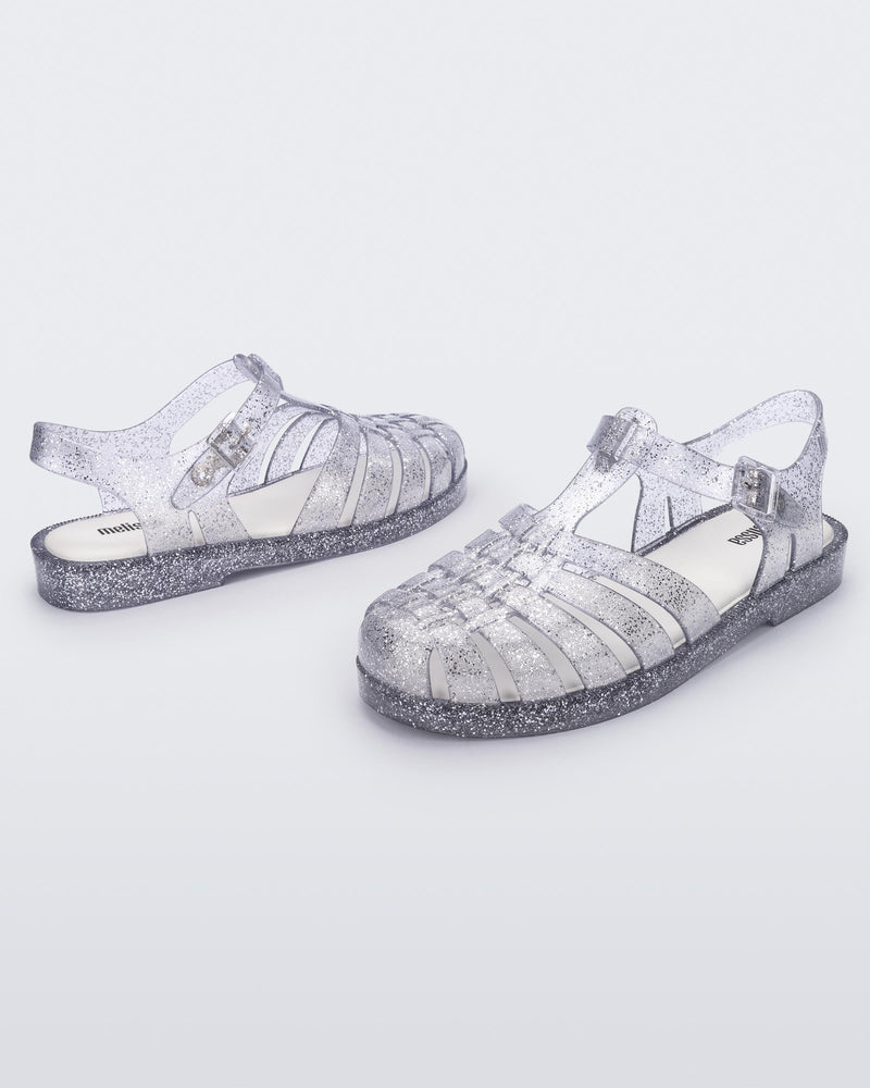 An angled front and side view of a pair of glitter clear Melissa Possession sandals with several straps and a closed toe front.