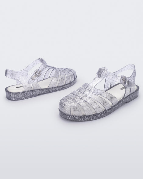 An angled front and side view of a pair of glitter clear Melissa Possession sandals with several straps and a closed toe front.