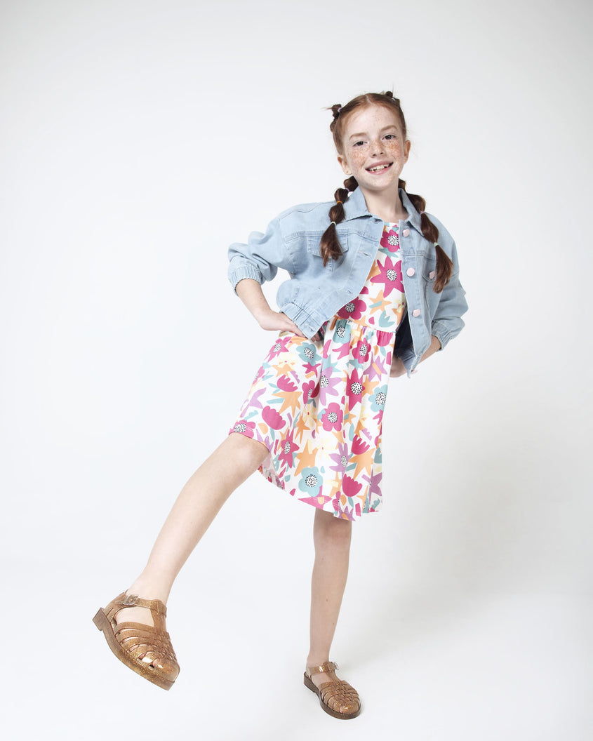 A model in a denim jacket, white patterned dress and a pair of Beige/Glitter/Gold Mini Melissa Possession Shiny sandals with several straps and a gold glitter base.