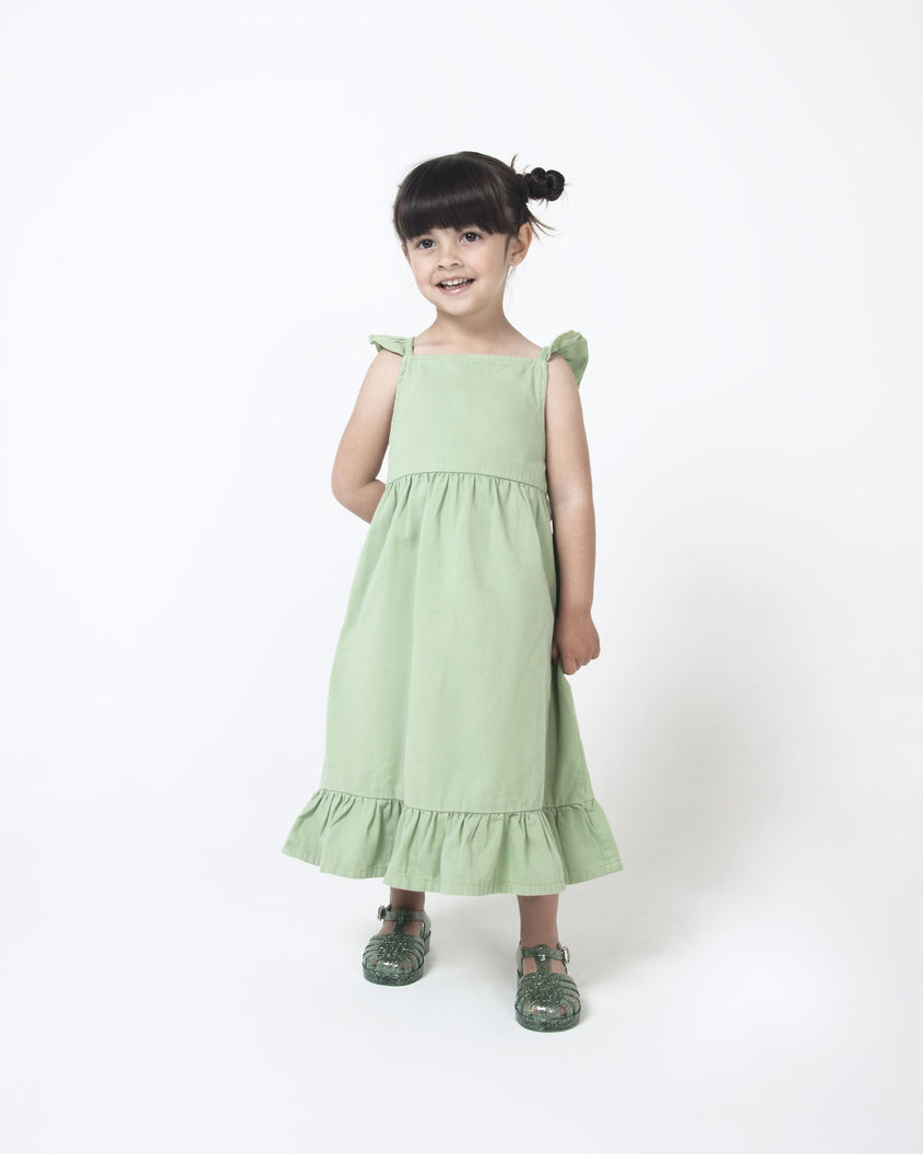 A young model in a green dress and a pair of Green/Glitter/Silver Mini Melissa Possession Shiny sandals with several straps, closed toe front and an ankle strap.